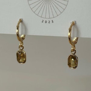 May include: Gold-toned hoop earrings with a rectangular, light-yellow gemstone pendant. The earrings are displayed against a white background with the text "BIG LITTLE DESIGN CO" and the year "2023".
