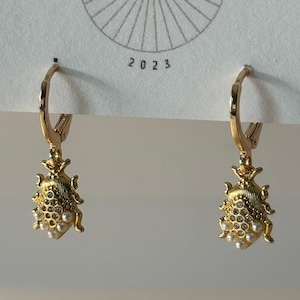 May include: A pair of gold-colored earrings with a lever back closure. Each earring features a detailed, bug-shaped charm adorned with small, faux pearls and clear crystals. The earrings hang from a white card with the text "DESIGN CO" and "2023".