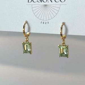 May include: Gold hoop earrings with rectangular light green faceted stones hanging from each hoop. The earrings are attached to a white display card featuring the text BIG LITTLE DESIGN CO and the year 2023 printed in black ink at the top.