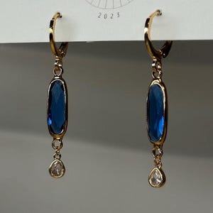 May include: Gold-tone hoop earrings with a blue, oval-shaped gemstone and a small, clear, teardrop-shaped stone. The earrings are hanging from a white card with the text "BIG LITTLE DESIGN CO" and the year "2023".
