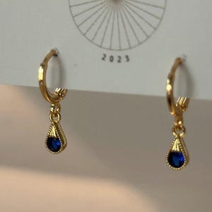 May include: A pair of gold-colored hoop earrings with a teardrop-shaped blue gemstone pendant. The earrings are displayed on a white card with the text "BIG LITTLE DESIGN CO" and the year "2023".