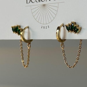 May include: Gold-colored hoop earrings with a cluster of green gemstones and a delicate chain. The earrings are displayed on a white card with the text "BIG LITTLE DESIGN CO." and the year "2023".