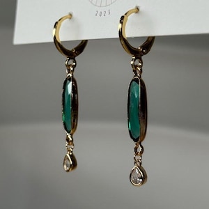 May include: Gold-tone hoop earrings with emerald green rectangular pendants and small teardrop-shaped clear stones. The earrings are displayed on a white card with the text "BIG LITTLE DESIGN CO" and the year "2023".