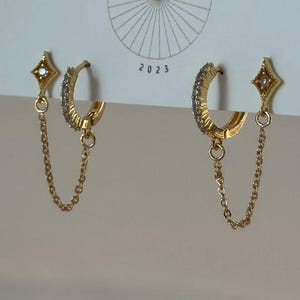 May include: A pair of gold-colored earrings with a chain and a small diamond-shaped charm. The earrings are displayed on a white card with the text "JEWELRY & THINGS" and "BIG LITTLE DESIGN CO 2023".
