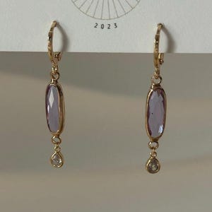 May include: A pair of gold-colored hoop earrings. Each earring features a light purple, oval-shaped stone and a small, clear teardrop-shaped stone. The earrings are displayed against a white background with the text "DESIGN CO" and the year "2023".
