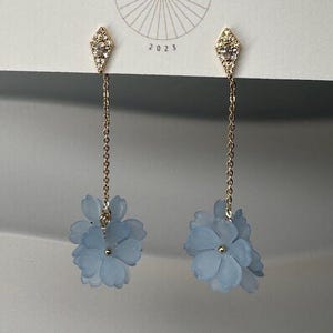 May include: A pair of gold-toned earrings with a diamond-shaped top encrusted with small clear stones. Delicate gold chains suspend light blue flower charms. The earrings are displayed on a white card with the text "BIG LITTLE DESIGN CO" and "2023".