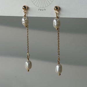 May include: A pair of gold-colored earrings with a pearl and chain design. The earrings have a small gold ball at the top, a pearl, a gold chain, and another pearl at the bottom. The earrings are hanging from a white card with the text "BIG LITTLE DESIGN CO" and the year "2023".