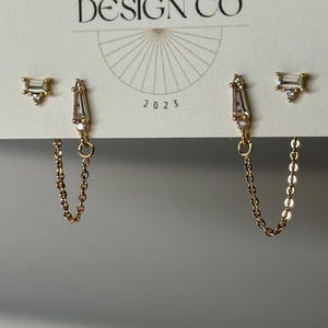 May include: Gold-toned earrings with clear, rectangular and triangular-shaped stones. One pair features a chain connecting two earrings. The card behind the earrings reads "BIG LITTLE DESIGN CO" and "2023".