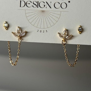 May include: A set of gold-colored earrings with clear, sparkling accents. One pair features a floral design with a dangling chain, while the other two are small studs. The earrings are displayed on a white card with the text "BIG LITTLE DESIGN CO."