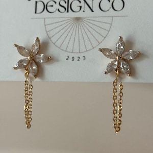 May include: A pair of gold-colored flower earrings with clear crystal accents and dangling chains. The earrings are displayed on a white card with the text "BIG LITTLE DESIGN CO" and the year "2023".