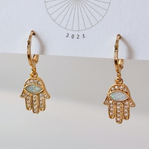 May include: A pair of gold-toned hoop earrings featuring dangling hamsa hand charms. Each charm is embellished with small clear stones and a central oval-shaped iridescent stone. The earrings are attached to a white display card marked with 2023.