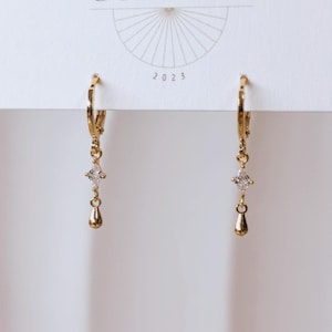 May include: A pair of gold-toned hoop earrings featuring a dangling marquise-cut clear stone and a small gold teardrop charm. The earrings are attached to a white display card with a fan-shaped graphic and the number 2023 printed on it.