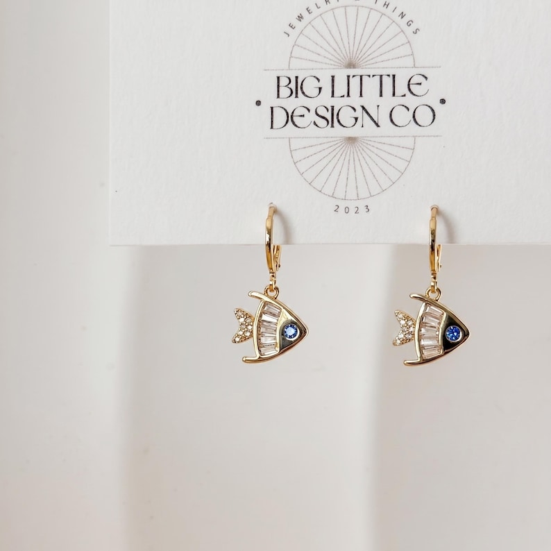 May include: Gold-tone fish earrings with a blue gem eye and clear crystal accents. The earrings are on a white card with the text "BIG LITTLE DESIGN CO" and "2023". The earrings have a hoop closure.