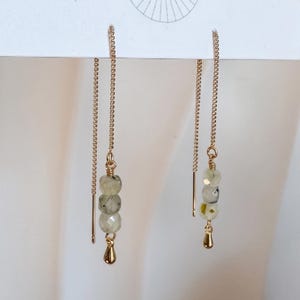 May include: A pair of gold-tone threader earrings featuring a delicate box chain. Each earring is adorned with three faceted, pale green, semi-translucent beads with dark inclusions, finished with a small, smooth gold-tone teardrop charm.