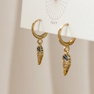 May include: A pair of gold-coloured hoop earrings with a feather-shaped charm. The charms are detailed with small blue and clear stones. The earrings are displayed on a white card with the year 2023 printed on it.