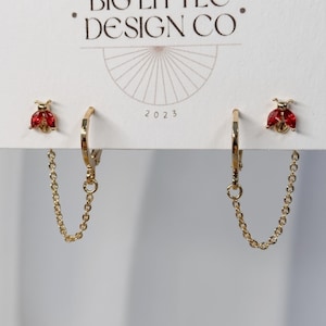 May include: Gold-tone hoop earrings with a delicate chain connecting the hoop to a ladybug stud. The ladybug features a vibrant red marquise-cut stone. The earrings are displayed on a white card labeled BIG LITTEC DESIGN CO 2023.