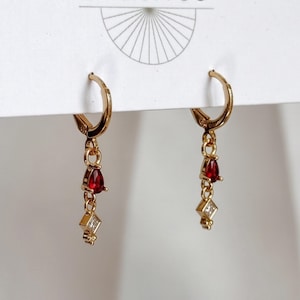 May include: A pair of gold hoop earrings featuring dangling charms. Each earring has a small red teardrop-shaped stone followed by a tiny square-shaped clear stone. The earrings are attached to a white display card with a partial sunburst graphic.