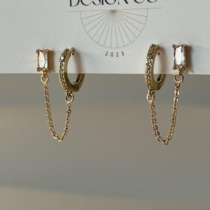 May include: A pair of gold-colored earrings with a rectangular clear stone and a small chain. One earring is a hoop style with small clear stones. The earrings are displayed on a white card with the text "BIG LITTLE DESIGN CO" and "2023".