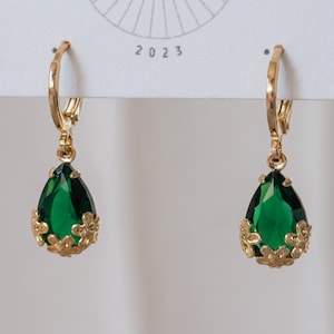 May include: A pair of gold-tone leverback earrings featuring faceted, pear-shaped emerald green stones. Each stone is encased at the base by a decorative gold-tone metal setting shaped like small flowers. The earrings are attached to a white card.