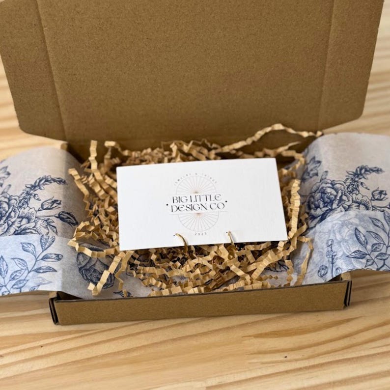 May include: An open cardboard box containing a white card with the text "BIG LITTLE DESIGN CO" and decorative floral tissue paper. The box is filled with brown crinkle paper. The tissue paper has a blue floral design.
