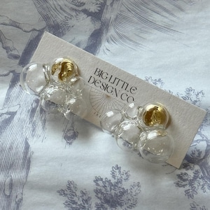 May include: A pair of gold and clear glass bubble earrings. The earrings are shaped like a cluster of bubbles and have a gold accent on each bubble. The earrings are on a white background with the text "BIG LITTLE DESIGN CO" printed on the background.