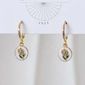 May include: A pair of gold-colored hoop earrings. Each earring features a small, round pendant with a white background and a painted tulip design in gold and green. The earrings are displayed against a white background with the year 2023 printed on the top.