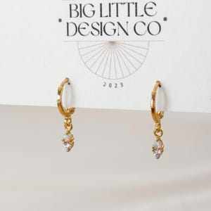 May include: A pair of gold hoop earrings attached to a white display card labeled BIG LITTLE DESIGN CO 2023. Each earring features a small dangling charm with a round iridescent opal stone positioned above a clear, faceted, round gemstone.