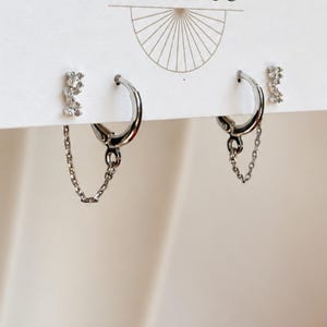 May include: A pair of silver-toned hoop earrings with attached delicate chains and two separate stud earrings featuring small clear stones. The jewelry is displayed on a white card with a thin line drawing of a sunburst design at the top.
