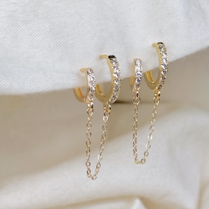 May include: Gold-toned hoop earrings with a delicate chain and sparkling clear stones. The earrings are set against a soft, white background, showcasing their elegant design. The earrings are a stylish accessory.