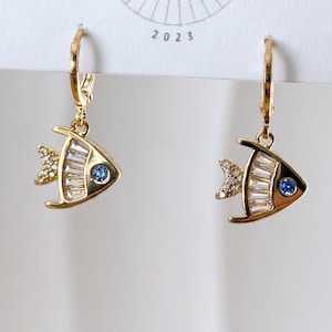 May include: Gold-toned fish-shaped earrings. Each earring features a fish design with a blue gem eye and a tail and body embellished with clear and sparkling crystals. The earrings are suspended from gold hoops.