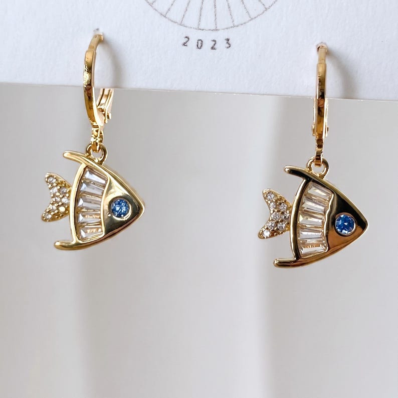 May include: Gold-toned fish-shaped earrings. Each earring features a fish design with a blue gem eye and a tail and body embellished with clear and sparkling crystals. The earrings are suspended from gold hoops.