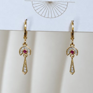 May include: A pair of gold-toned drop earrings featuring a semi-circular top section encrusted with small clear stones and a central round pink stone. A slender, pointed, stone-set dangle hangs from the center of each semi-circle.