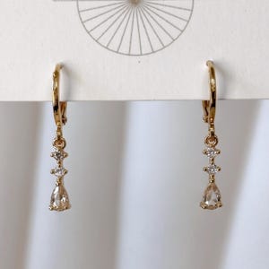 May include: A pair of gold-toned hoop earrings featuring a vertical drop design. Each earring consists of two small round clear stones followed by a single pear-shaped clear stone dangling from a hinged hoop closure attached to a white display card.