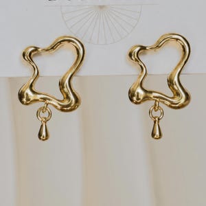 May include: A pair of gold-toned earrings featuring an irregular, open-form organic shape. Each earring includes a small, smooth teardrop-shaped charm suspended from the bottom of the frame by a circular jump ring, creating a dangling effect.