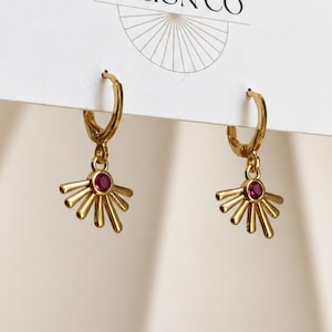 May include: A pair of gold hoop earrings featuring a dangling sunburst charm. Each charm is centered with a small, round, deep pink gemstone. The earrings are attached to a white display card with a partial sunburst logo and text at the top.