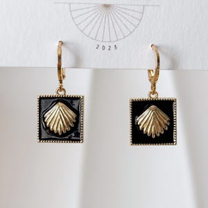 May include: Gold-tone hoop earrings with black square pendants. Each pendant features a gold seashell design. The earrings are displayed against a white background with the year 2023 printed at the top.