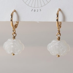 May include: A pair of gold-toned hoop earrings featuring dangling white, cloud-shaped carved stone charms. Each charm is secured with a small gold-toned bead at the base and attached to the hoop with a gold-toned wire wrap, suspended from a white card.