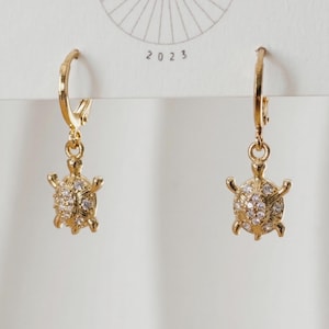 May include: A pair of gold-colored hoop earrings. Each earring features a small, detailed turtle pendant encrusted with clear gemstones. The earrings are displayed against a white background, with the year "2023" visible.