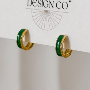 May include: A pair of small gold hoop earrings featuring a vibrant green enamel inlay. The earrings are attached to a white display card that has the text DESIGN CO printed at the top with a semi-circular line graphic underneath the text.