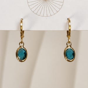 May include: A pair of gold-toned leverback earrings featuring oval-shaped, faceted deep blue-green stones set in polished metal bezels. The earrings hang from a white display card that features a thin, circular line graphic at the top center.