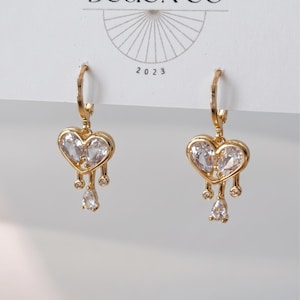 May include: A pair of gold-tone hoop earrings featuring a heart-shaped charm with a clear, faceted stone center. The heart design includes three dangling accents: two small circular stones and one central pear-shaped stone, all in a gold setting.