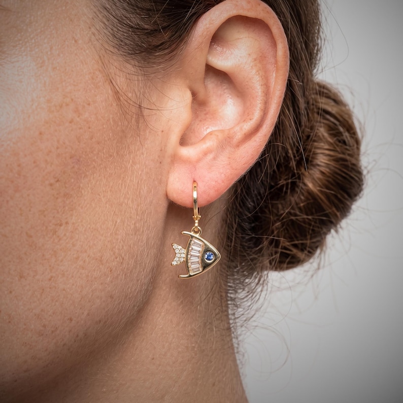 May include: Gold fish-shaped earring with a blue gem eye and white baguette-cut stones. The earring is on a gold hoop. The fish has a small fin with white stones. The earring is hanging from a person's ear.