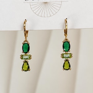 May include: A pair of gold-tone drop earrings featuring a vertical arrangement of three faceted stones: a dark green oval at the top, a light green rectangular baguette in the center, and a pear-shaped olive green stone at the bottom.