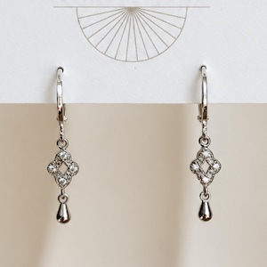 May include: A pair of silver-toned leverback earrings featuring a dangling quatrefoil charm encrusted with small clear stones, finished with a smooth, polished teardrop-shaped drop at the bottom, displayed on a white card with a fan graphic.
