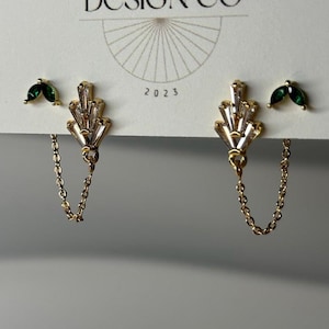 May include: A pair of gold-toned earrings with clear, baguette-cut crystals and a delicate chain. The earrings are paired with small, green leaf-shaped studs. The card behind the earrings reads "BIG LITTLE DESIGN CO" and "2023".