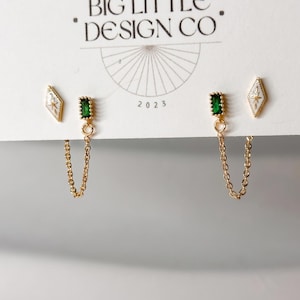 May include: A pair of gold-tone earrings on a display card labeled BIG LITTLE DESIGN CO. Each earring features a white diamond-shaped stud with a star detail and a rectangular green stone stud connected by a delicate gold chain to the ear post.
