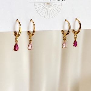 May include: Two pairs of gold hoop earrings attached to a white display card. Each hoop features a small, dangling teardrop-shaped gemstone. The stones vary in color, including deep red and light pink hues, suspended from polished gold metal hoops.