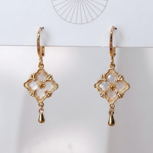 May include: A pair of gold-toned drop earrings featuring a four-pointed clover-shaped frame with a white mother-of-pearl inlay. Each earring has a small central gold flower detail and a tiny dangling teardrop charm suspended from the bottom edge.
