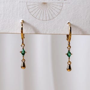 May include: A pair of gold hoop earrings featuring a dangling design. Each earring consists of a small gold hoop, a central green diamond-shaped gemstone, and a smooth gold teardrop-shaped charm at the bottom, suspended from a white display card.