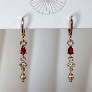 May include: A pair of gold-toned dangle earrings featuring a lever-back closure, a teardrop-shaped red stone, a small square-cut clear stone set in a diamond-shaped frame, and a round gold bead at the bottom of each earring, displayed on a white card.